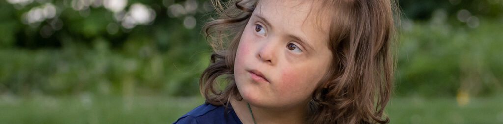 Ein junges Mädchen mit Down-Syndrom blickt versonnen erwartungsvoll schräg nach oben.